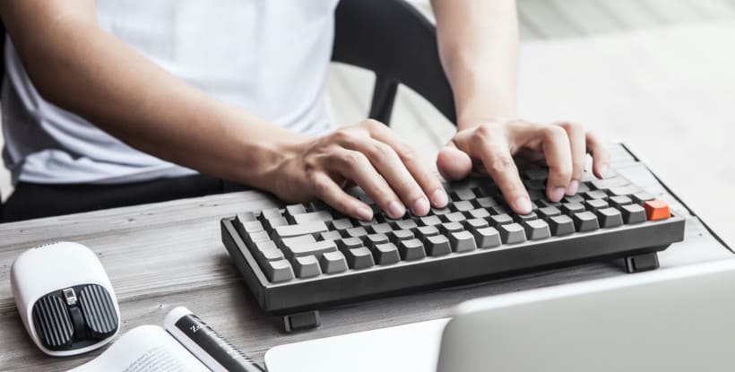 Copywriter zaprasza do kontaktu. Pisze teksty na stronę przy pomocy klawiatury, obok leży mysz i gazeta, widać ekran laptopa.